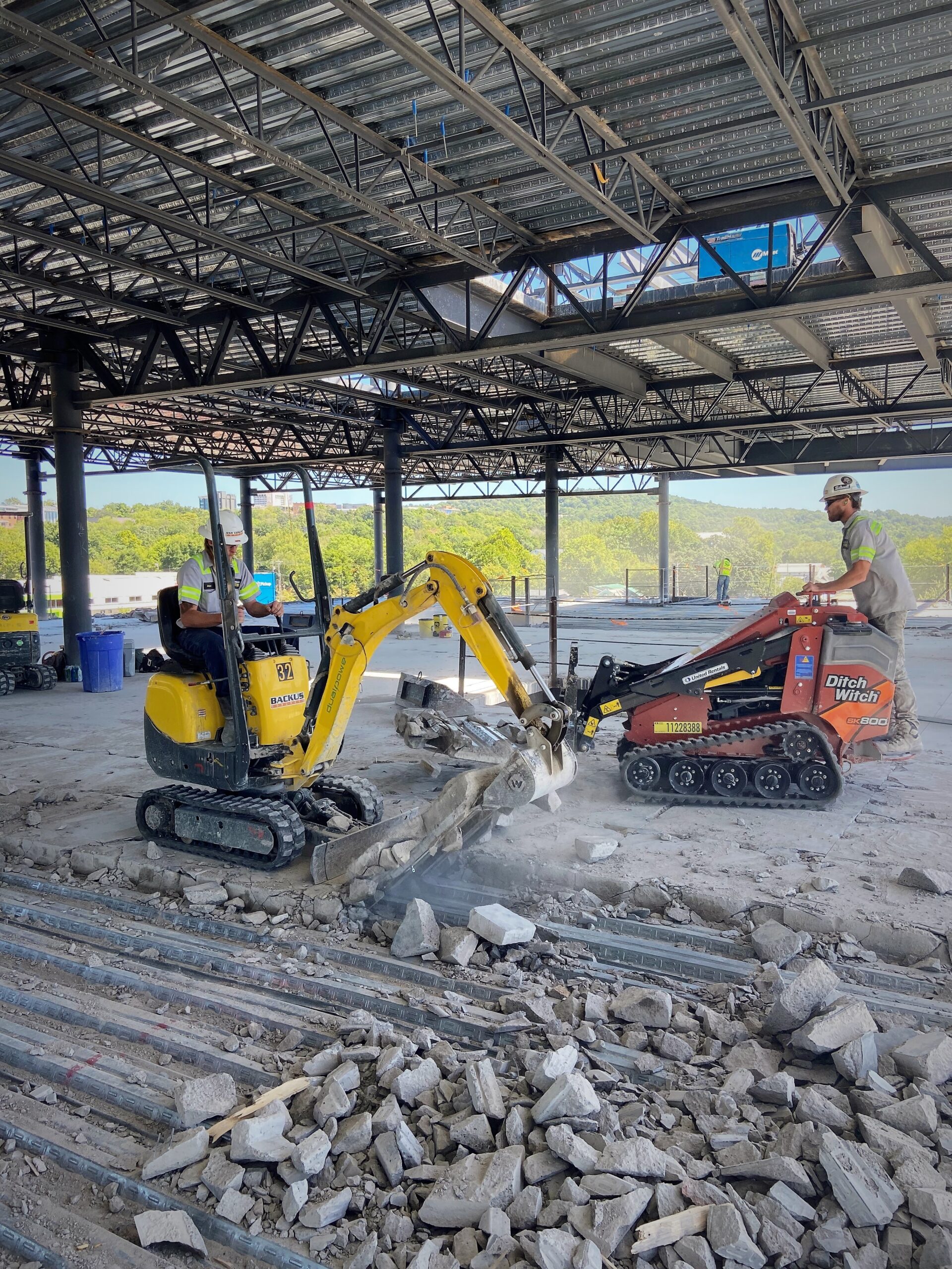 An Any Concrete Cutting contractor on an excavator doing concrete demolition work underneath a metal awning and the other technician on a skid steer